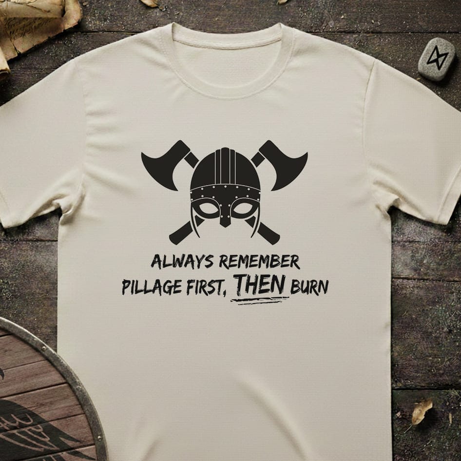 An off-white short-sleeve t-shirt with a black graphic of a Viking helmet and crossed axes, featuring the text 'ALWAYS REMEMBER PILLAGE FIRST, THEN BURN'. The shirt is laid flat on a dark rustic wooden background with Viking-themed props.
