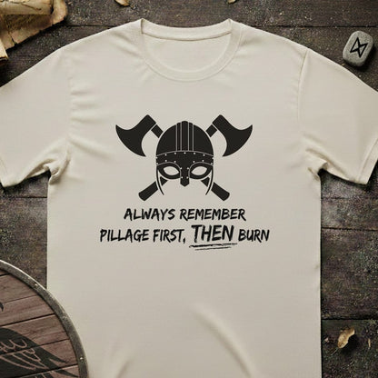 An off-white short-sleeve t-shirt with a black graphic of a Viking helmet and crossed axes, featuring the text 'ALWAYS REMEMBER PILLAGE FIRST, THEN BURN'. The shirt is laid flat on a dark rustic wooden background with Viking-themed props.