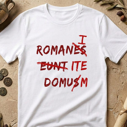 A white short-sleeve t-shirt with a red distressed text graphic reading 'I ROMANES CUNTI TE DOMUȘM' in Romanian, laid flat on a textured beige stone surface with decorative props like coins, dried leaves, and a rolled paper scroll.
