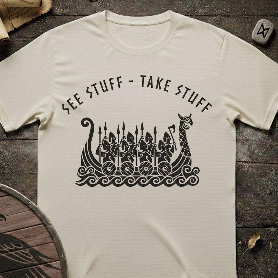 An off-white t-shirt laid flat on a dark rustic wooden surface, featuring a black graphic of a Viking longship with warriors and the text 'SEE STUFF - TAKE STUFF' arched above it.