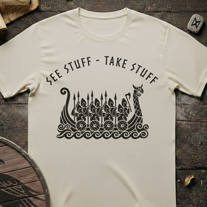 An off-white t-shirt laid flat on a dark rustic wooden surface, featuring a black graphic of a Viking longship with warriors and the text 'SEE STUFF - TAKE STUFF' arched above it.