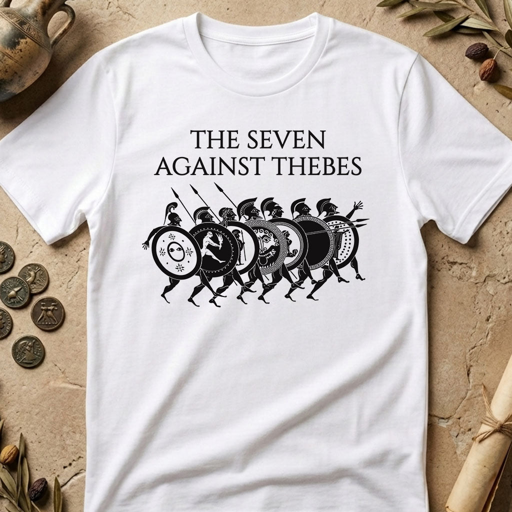 A white short-sleeve crewneck t-shirt with a black graphic print of seven ancient Greek-style warriors and the text 'THE SEVEN AGAINST THEBEES'. The shirt is displayed in a flat-lay arrangement on a textured beige stone surface with decorative props including coins, dried leaves, and a scroll.