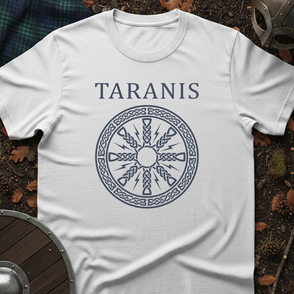 A white short-sleeve T-shirt laid flat on a dark forest floor with autumn leaves, showing a dark blue printed design of the word 'TARANIS' above a circular Celtic-style wheel motif. A shield, plaid fabric, and part of a helmet are visible around the edges as styled props.