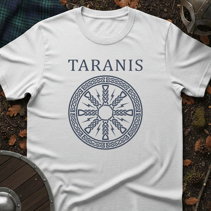 A white short-sleeve T-shirt laid flat on a dark forest floor with autumn leaves, showing a dark blue printed design of the word 'TARANIS' above a circular Celtic-style wheel motif. A shield, plaid fabric, and part of a helmet are visible around the edges as styled props.