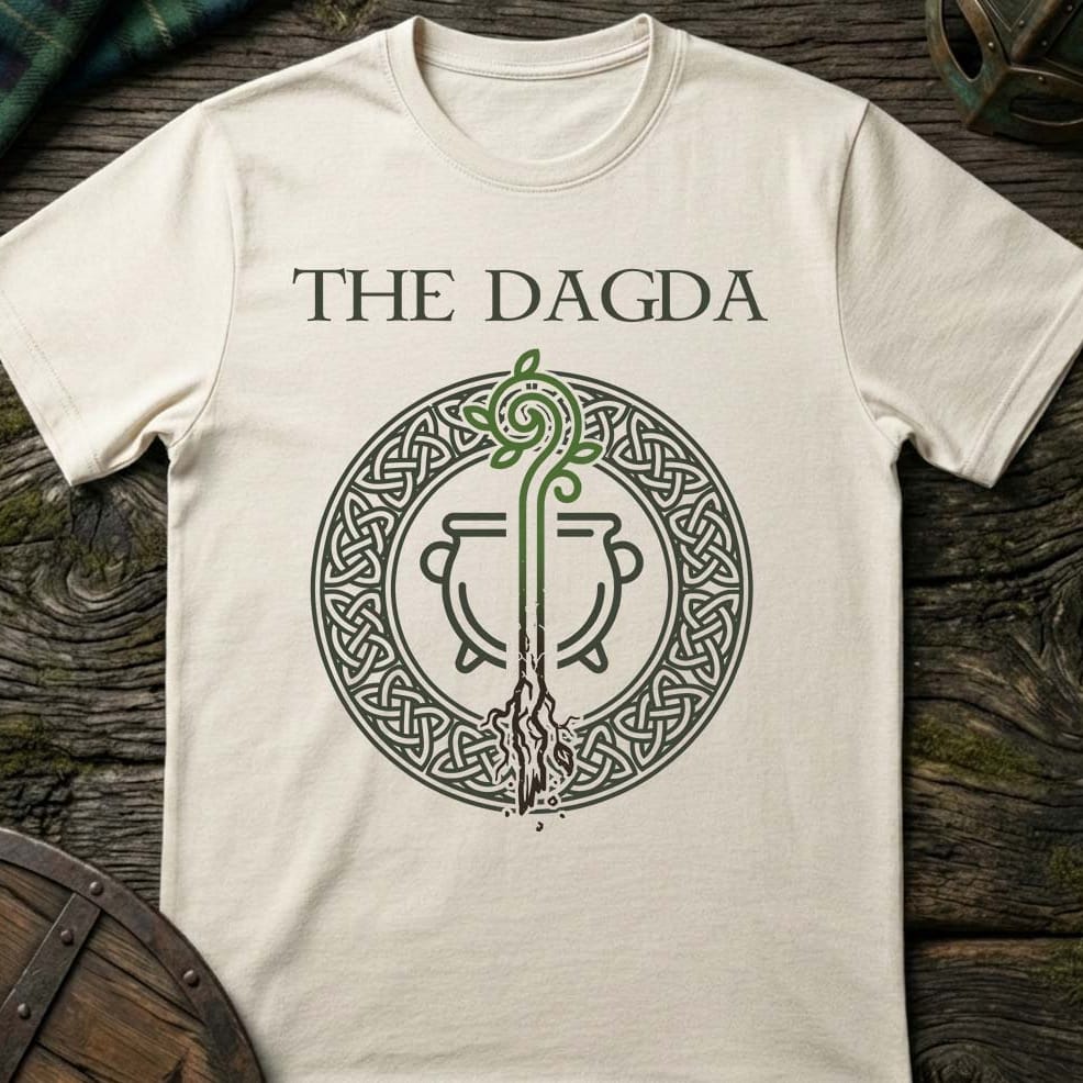 An off-white crewneck t-shirt featuring a dark green graphic of the 'The Dagda' cauldron emblem, laid flat on a rustic dark wood background.
