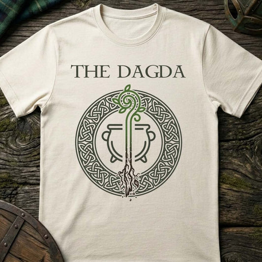 An off-white crewneck t-shirt featuring a dark green graphic of the 'The Dagda' cauldron emblem, laid flat on a rustic dark wood background.