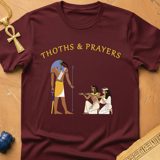 Maroon t-shirt with Egyptian-themed design and 'THOths & Prayers' text on a wooden surface.