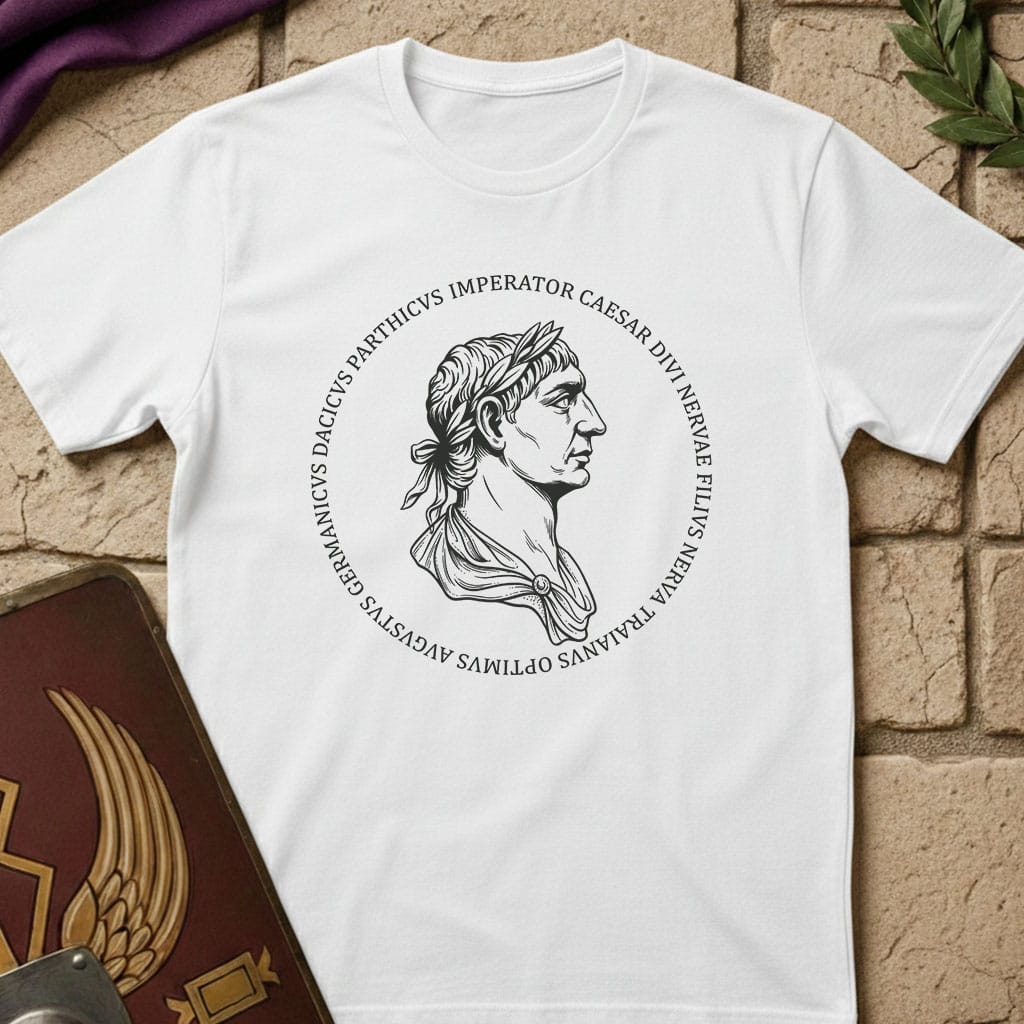 A white short-sleeve T-shirt laid flat on a tan stone-textured background, featuring a central graphic of a sword surrounded by green laurel branches and the words 'THE CATILINE CONSPIRACY' in black. Decorative props including a purple fabric corner, a green leaf, and part of a brown book are visible around the shirt.