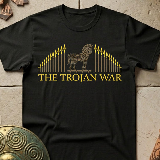A black short-sleeve crewneck t-shirt featuring a gold graphic of the Trojan Horse flanked by arrows and the text 'THE TROJAN WAR' in gold. The shirt is displayed flat on a textured stone surface with decorative bronze shields in the corners.