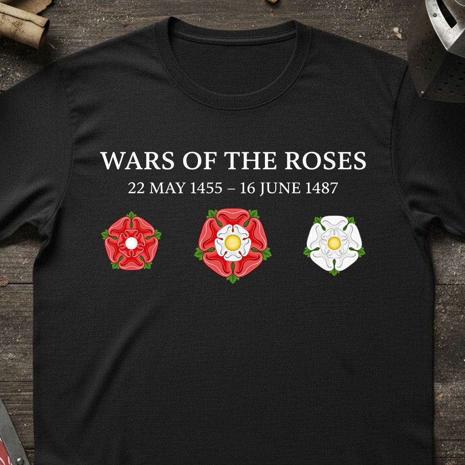 A black short-sleeve t-shirt laid flat on a dark wooden surface, featuring the text 'WARS OF THE ROSES 22 MAY 1455 - 16 JUNE 1487' above three rose emblems in red, white, and green. The shirt is styled with rustic props around the edges.