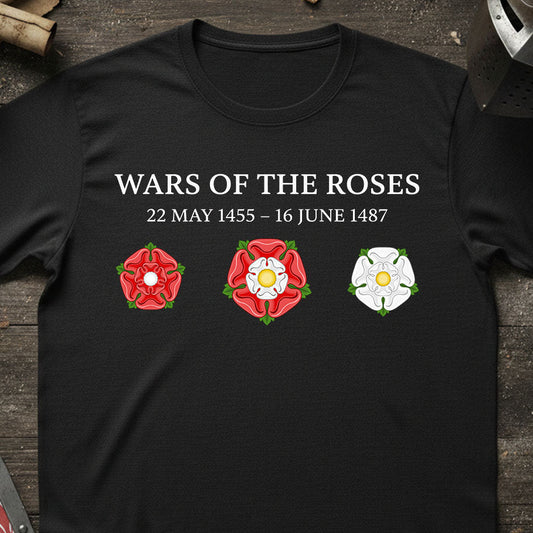 A black short-sleeve t-shirt laid flat on a dark wooden surface, featuring the text 'WARS OF THE ROSES 22 MAY 1455 - 16 JUNE 1487' above three rose emblems in red, white, and green. The shirt is styled with rustic props around the edges.